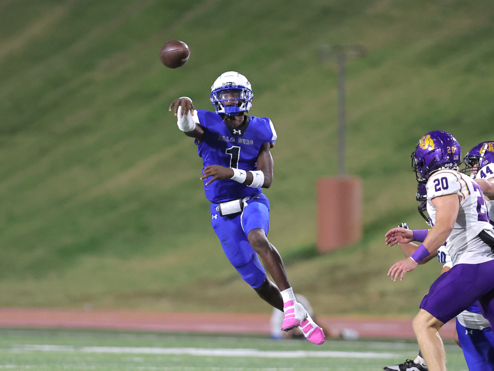 American football action: a blue-uniformed player reaches to catch a pass as the ball flies overhead, with purple-helmeted defenders nearby on a grassy field.