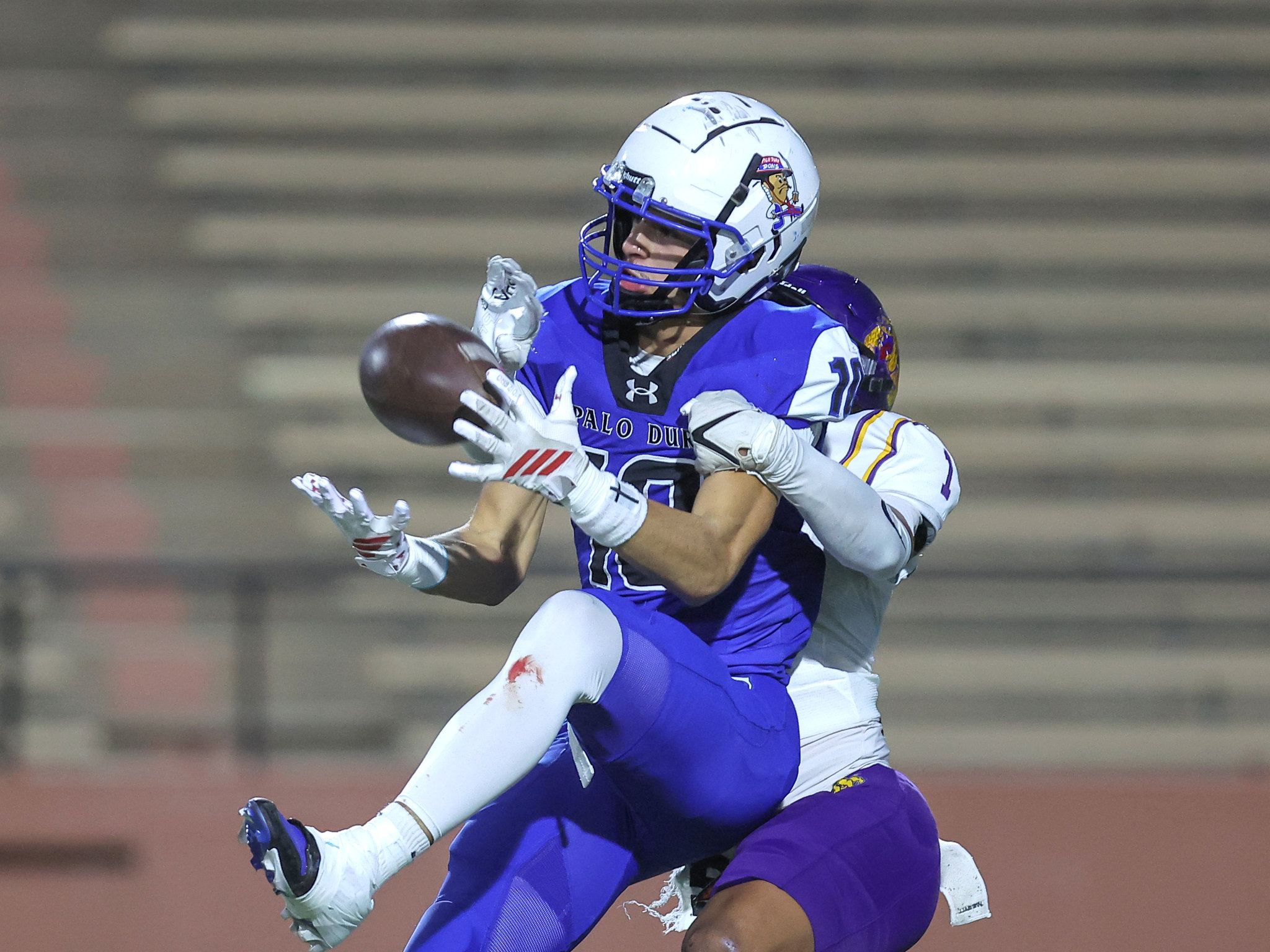 Two football players in purple uniforms jump for the ball during a game, reaching with gloved hands as one tries to catch it or intercept.