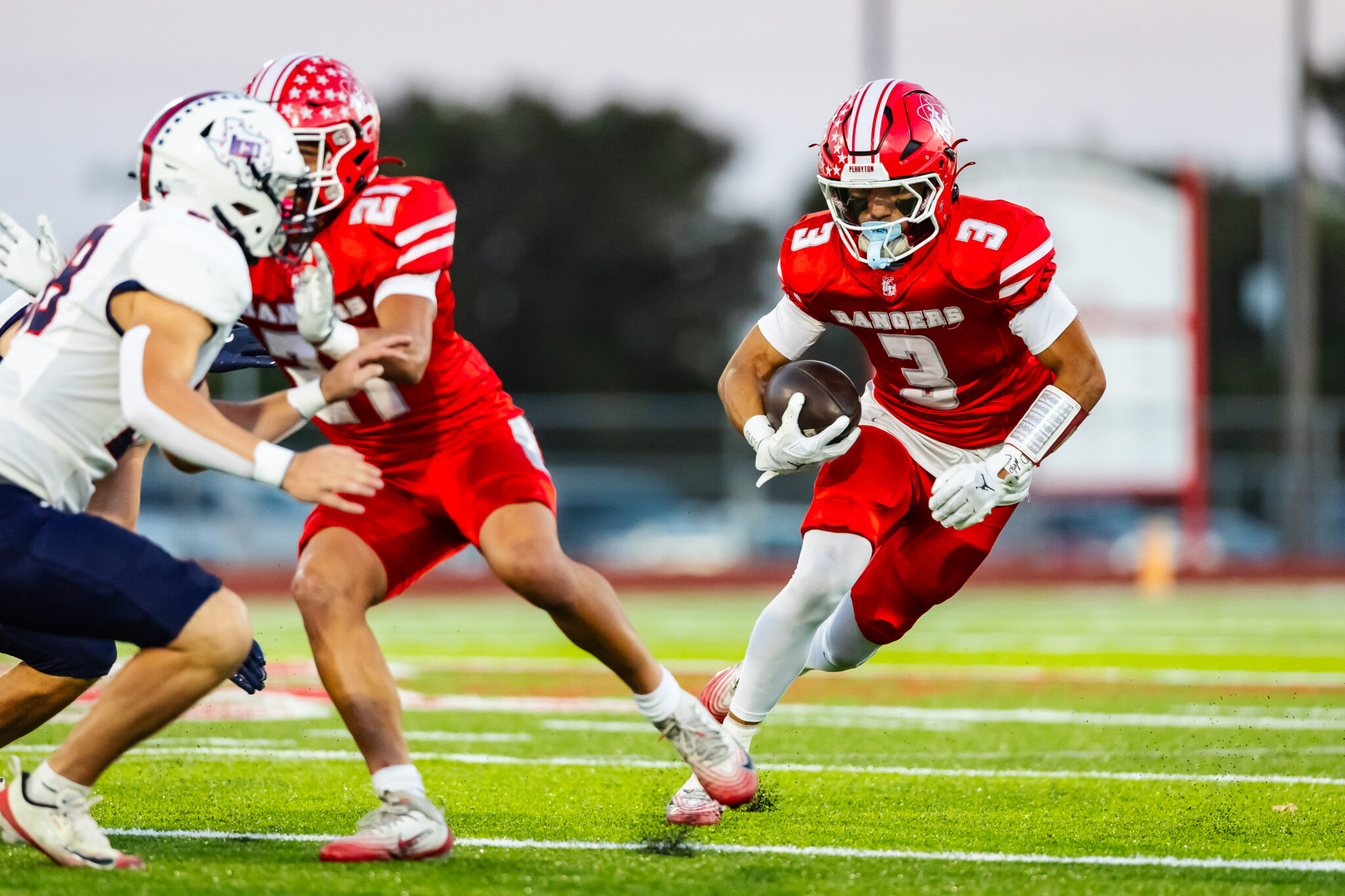 100A5646 | Press Pass Sports Football player in red uniform runs with the ball as defenders close in on a green field during a game, focused and sprinting forward.