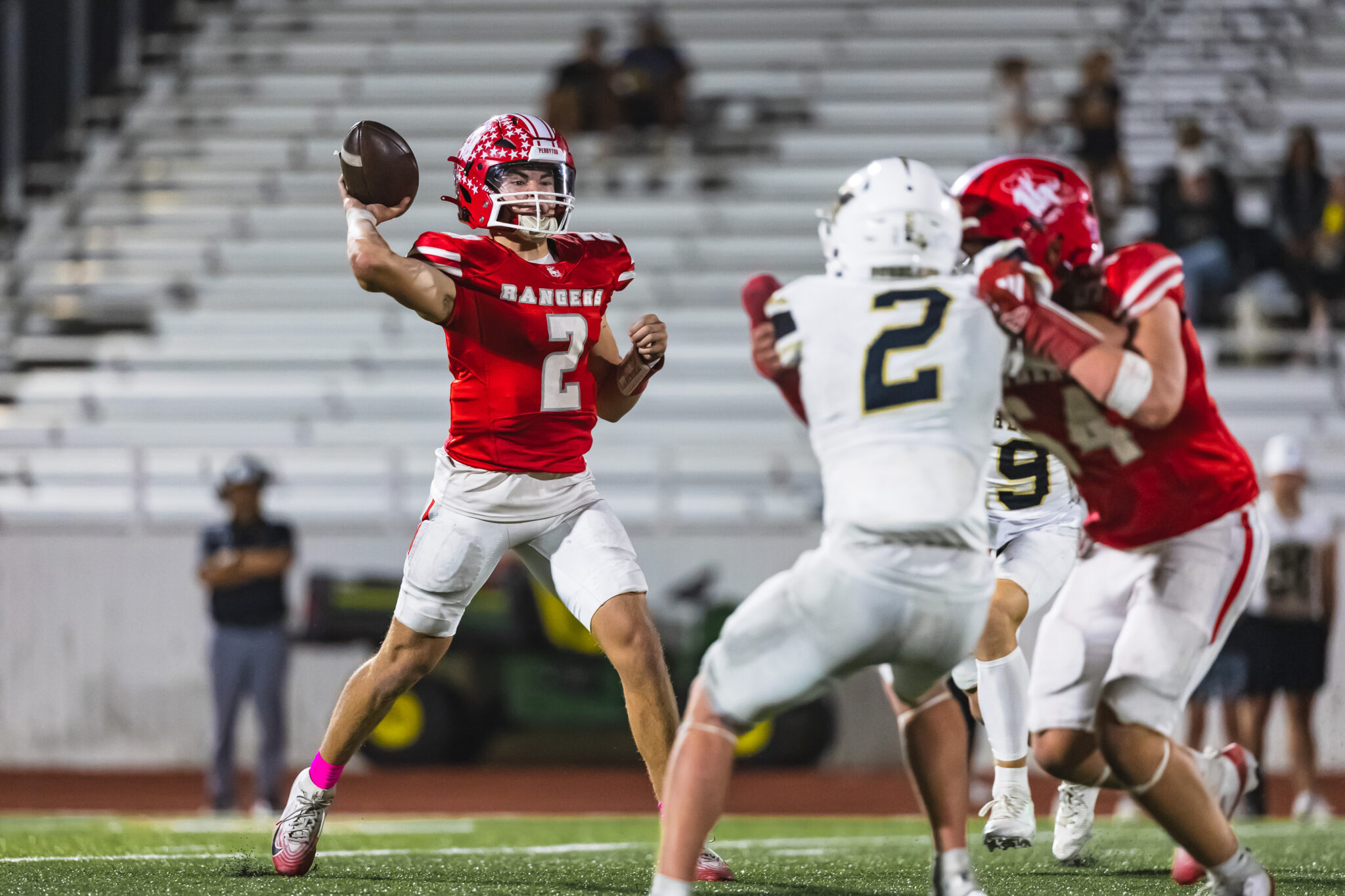 100A2025 1 | Press Pass Sports Quarterback in a red jersey throws a pass while teammates and defenders clash in the background on a football field.