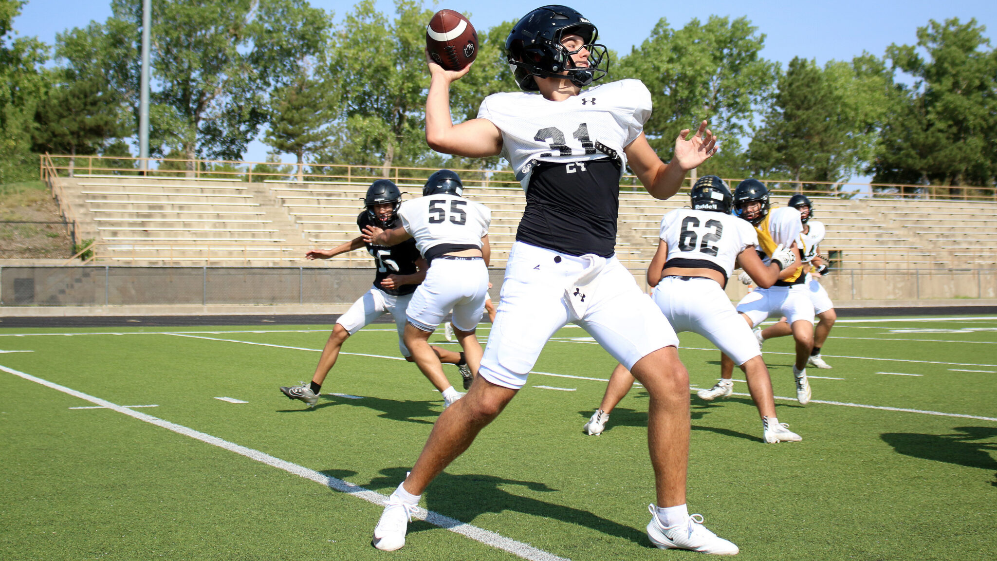 Canadian - Weston Mitchell 3 | Press Pass Sports Quarterback in white/black uniform throws a pass while teammates scramble on a sunny football field with bleachers in the background.