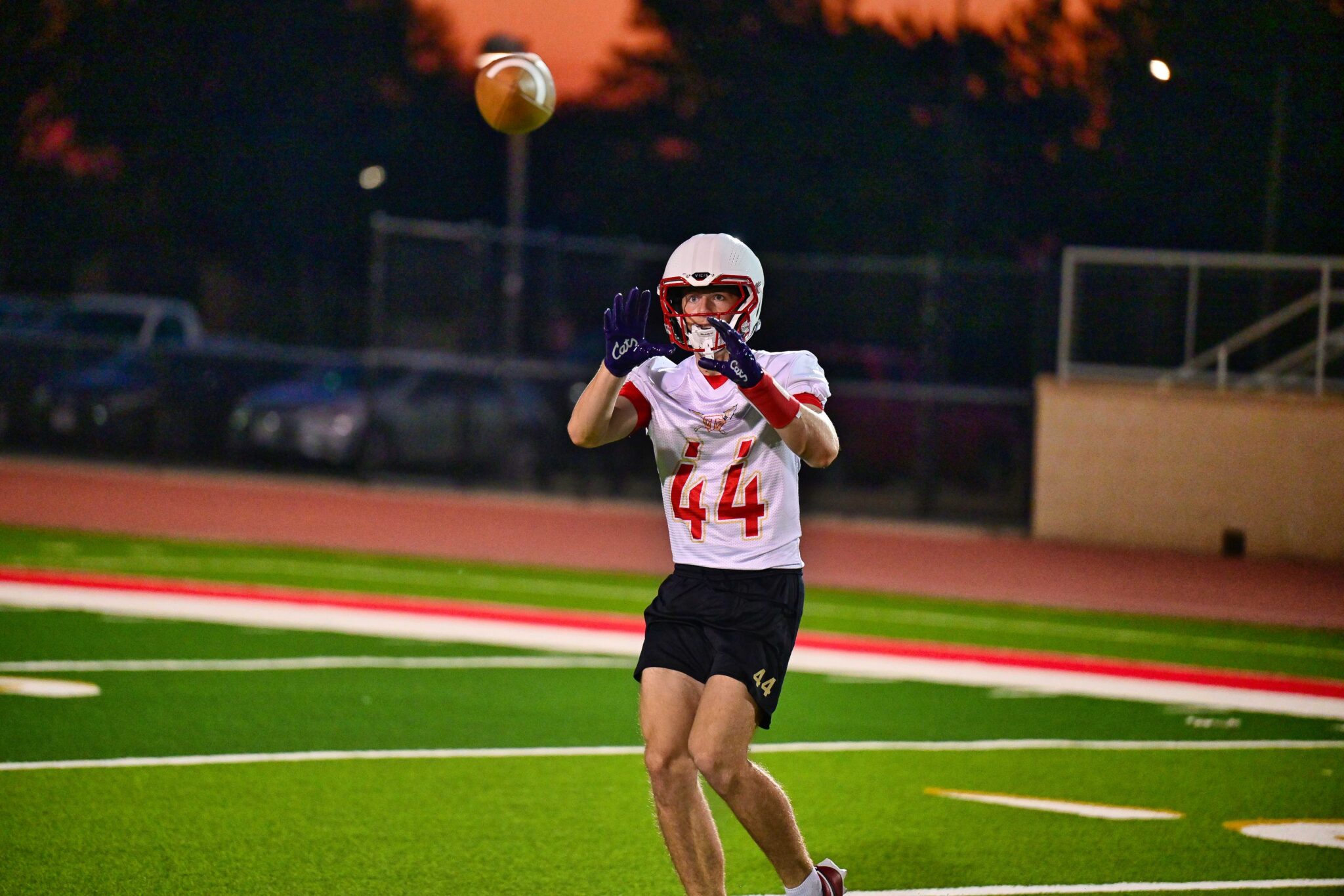 image6 6 | Press Pass Sports Football player in a white jersey and helmet reaching to catch a pass on a lit night field.