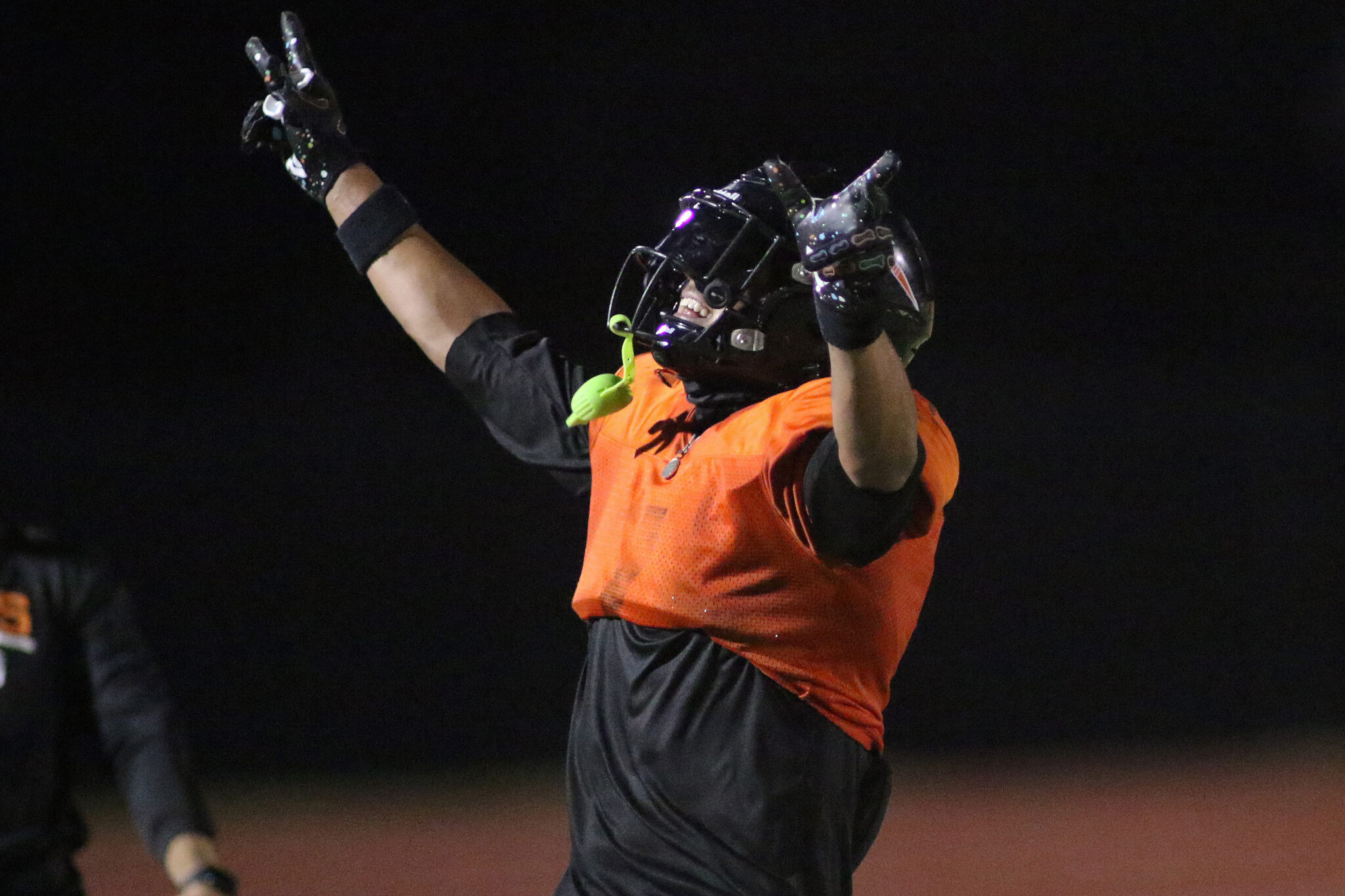 1K6A3031 | Press Pass Sports Athlete in an orange vest raises both gloved hands in celebration on a dark track at night, wearing a helmet.