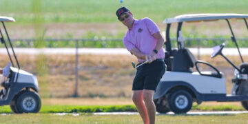 Golfer in a pink polo swinging a club on a sunny golf course with carts in the background, ball in the air