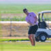 Golfer in a pink polo swinging a club on a sunny golf course with carts in the background, ball in the air