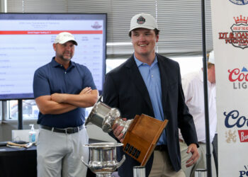 CoorTOC_FinalDay-72 | Press Pass Sports Young man in a blazer and cap smiling while holding a tall silver trophy at a golf tournament awards area; another man stands with arms crossed in the background.