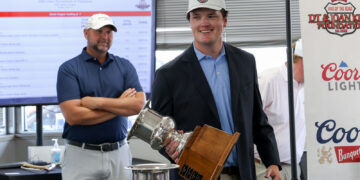 Young man in a blazer and cap smiling while holding a tall silver trophy at a golf tournament awards area; another man stands with arms crossed in the background.