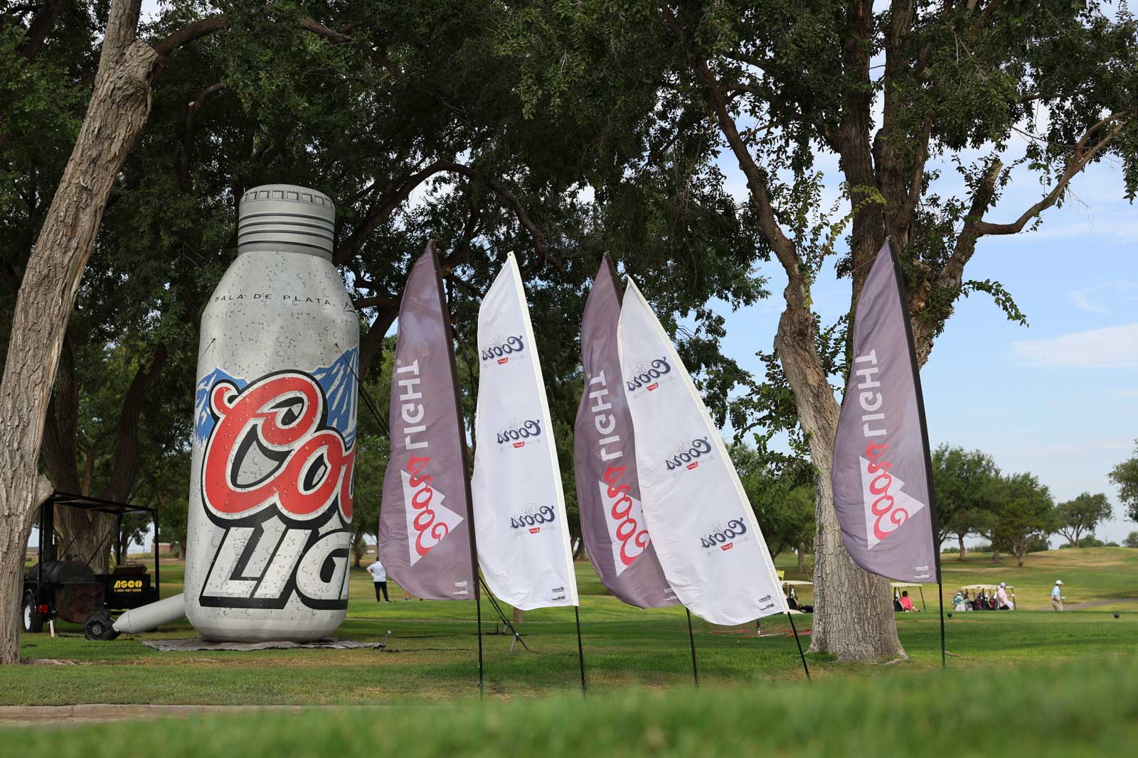 CoorTOC_FinalDay-2 | Press Pass Sports Large Coca‑Cola inflatable bottle with tall branded flags set up on a golf course, under trees on a sunny day.