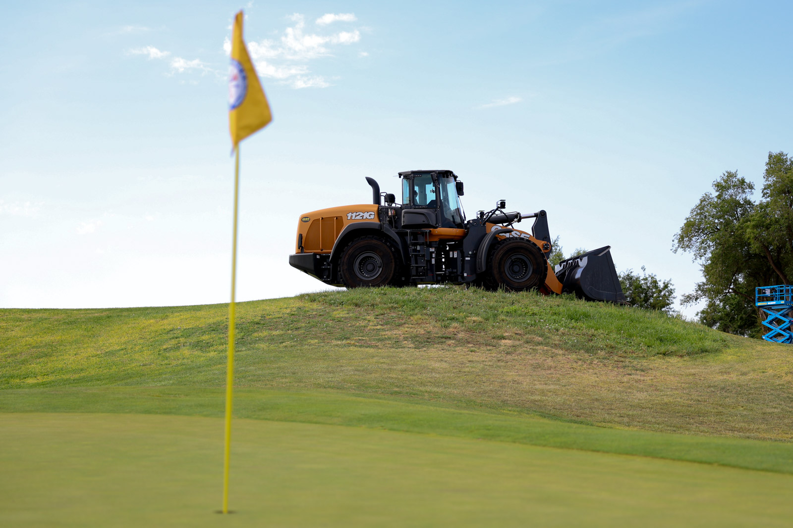 CoorTOC_FinalDay-5 | Press Pass Sports Orange heavy bulldozer with a large front bucket sits on a grassy hill, near a yellow flag on a golf green.