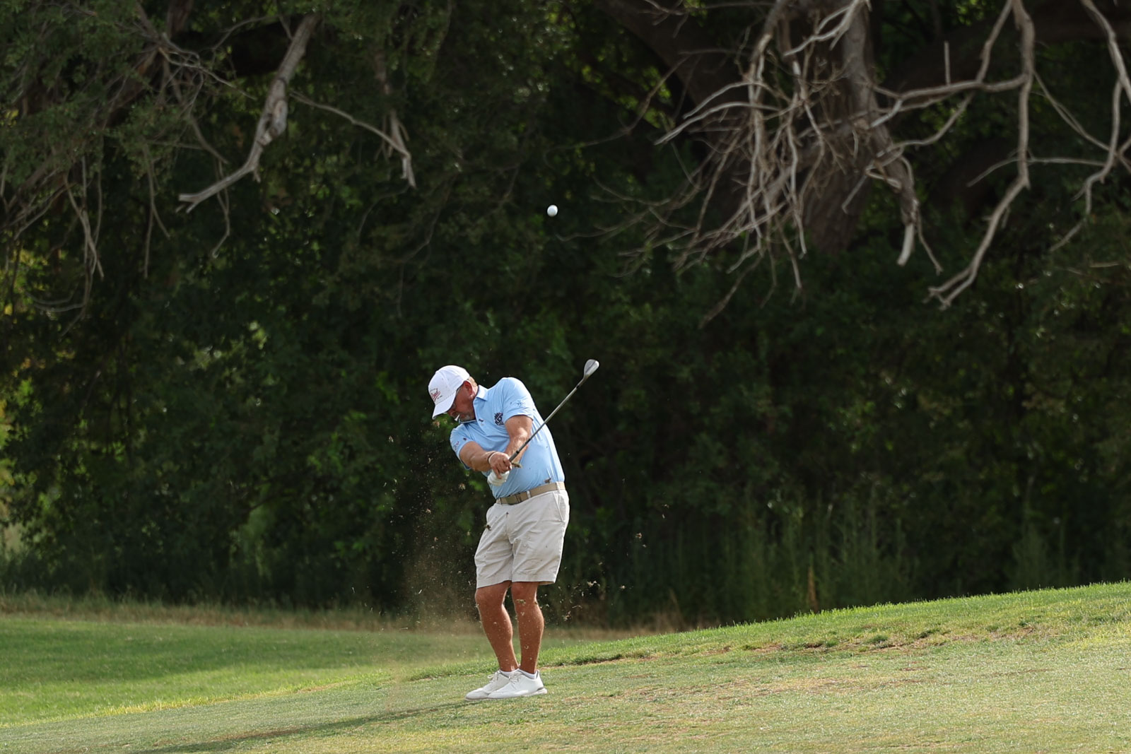CoorTOC_FinalDay-6 | Press Pass Sports Golfer in a light blue shirt and beige shorts mid-swing on a grassy fairway, ball airborne with trees behind him.