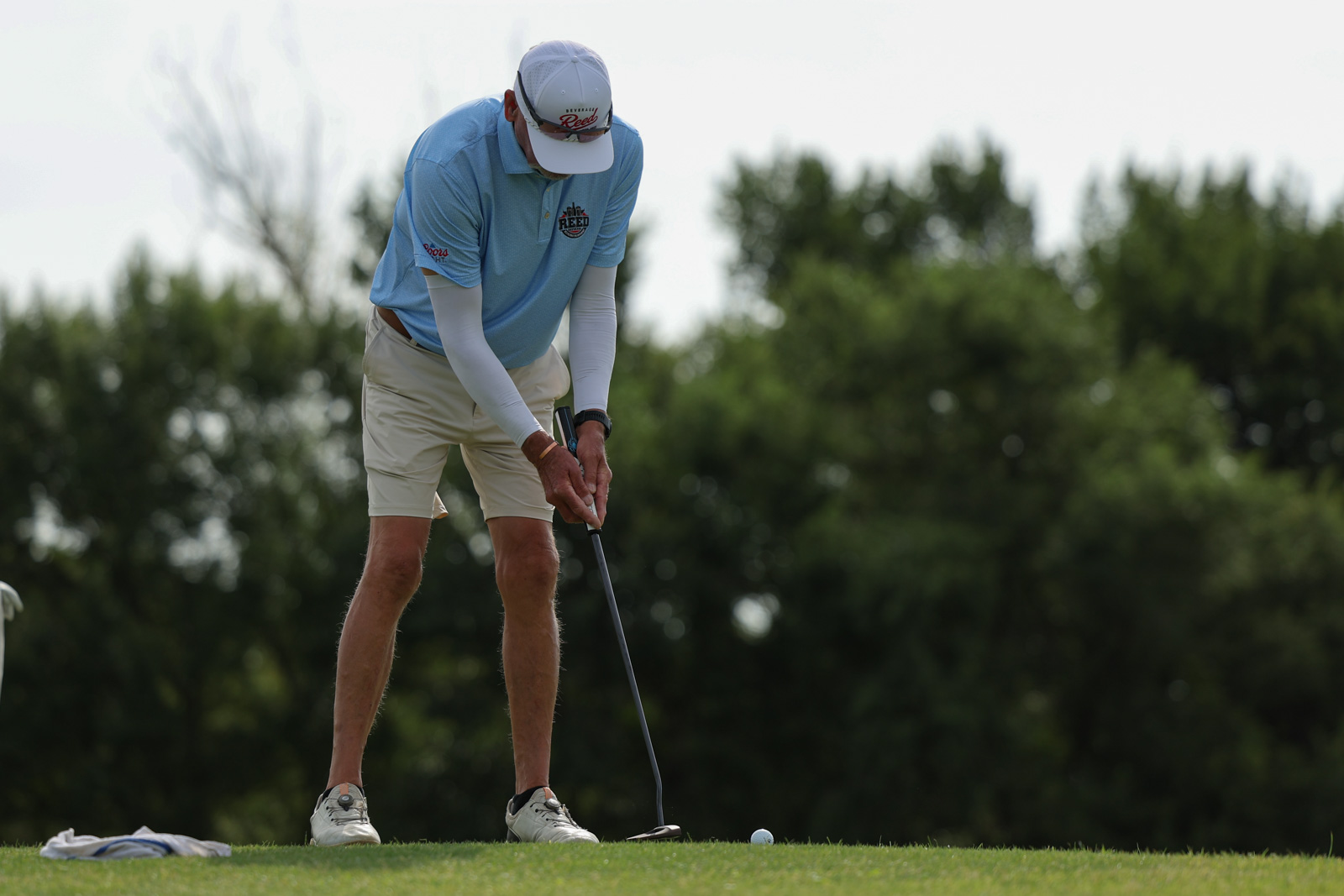 CoorTOC_FinalDay-11 | Press Pass Sports Golfer in a light blue polo and white cap lining up a putt on the green, with a golf ball nearby and trees in the background.