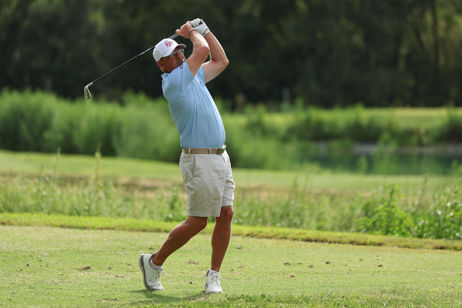 CoorTOC_FinalDay-17 | Press Pass Sports Male golfer in a light blue shirt, beige shorts, and white cap mid-swing on a sunny golf course. The green fairway and trees form the background.