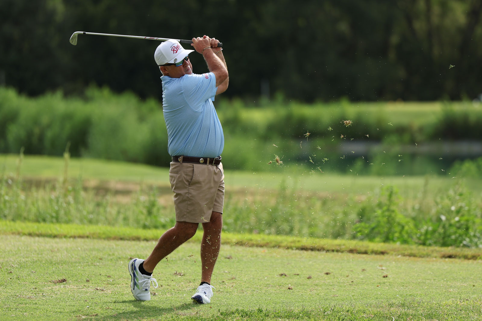 CoorTOC_FinalDay-19 | Press Pass Sports A man in a light blue polo and beige shorts swings a golf club on a lush course, dirt and grass bits flying from his shot.