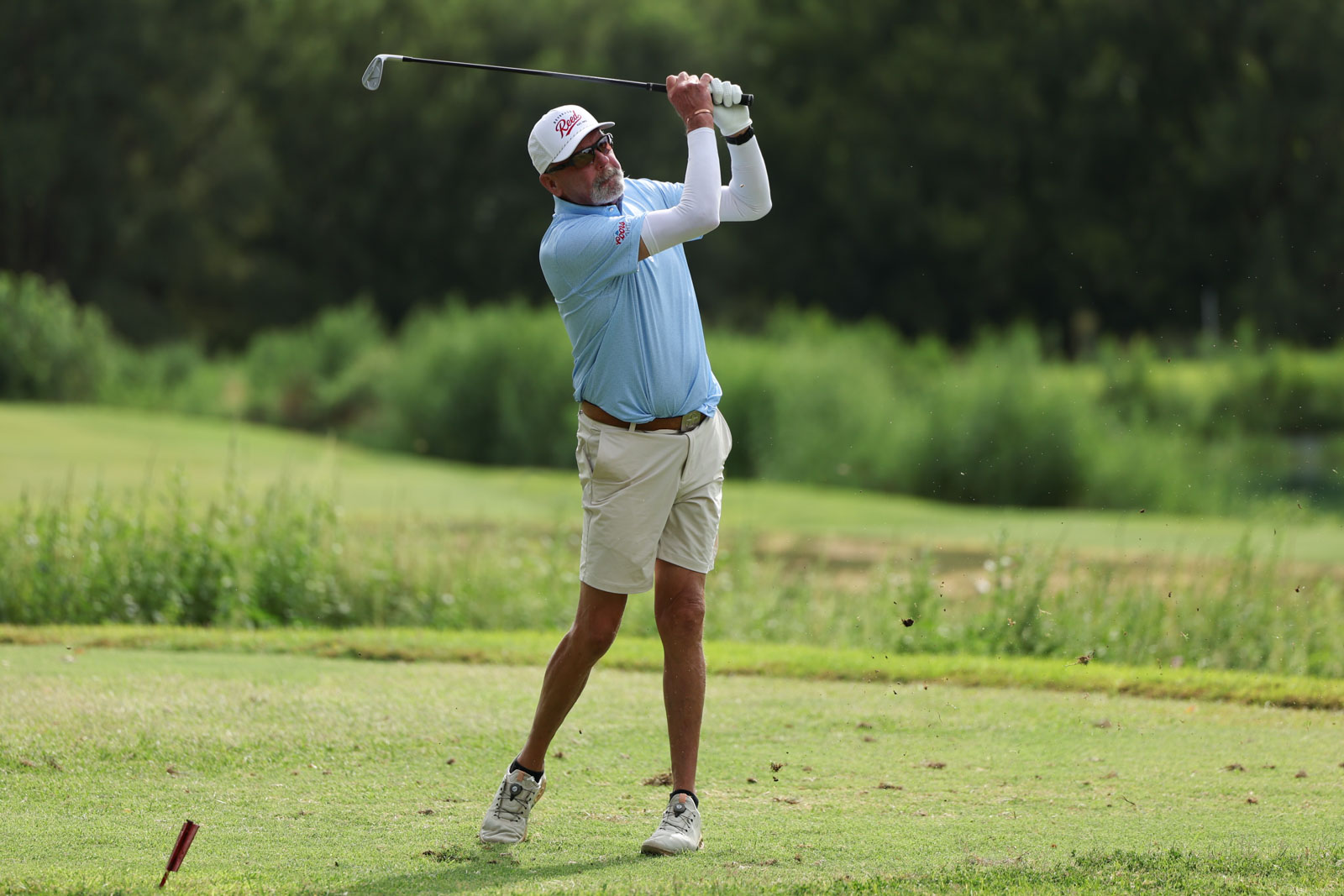 CoorTOC_FinalDay-20 | Press Pass Sports A man wearing a light blue shirt and beige shorts swings a golf club on a grassy golf course, mid-swing with a focused look.