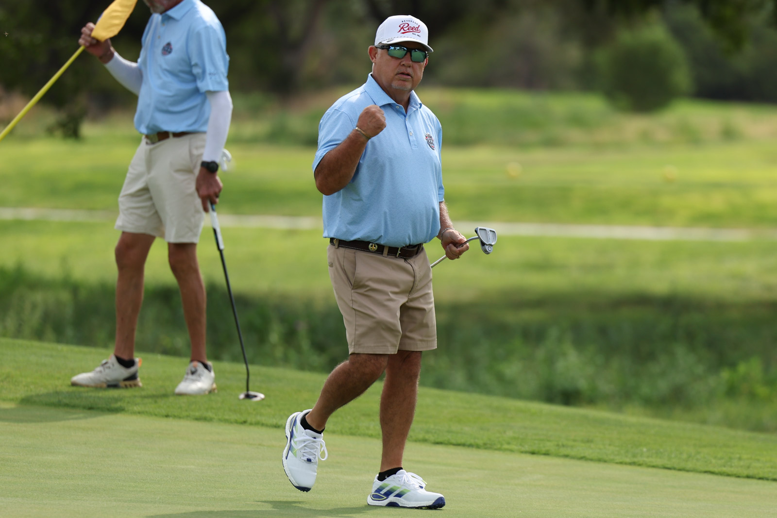 CoorTOC_FinalDay-22 | Press Pass Sports Man in light blue polo raises fist in celebration on a golf green, holding a club; another golfer stands in background.