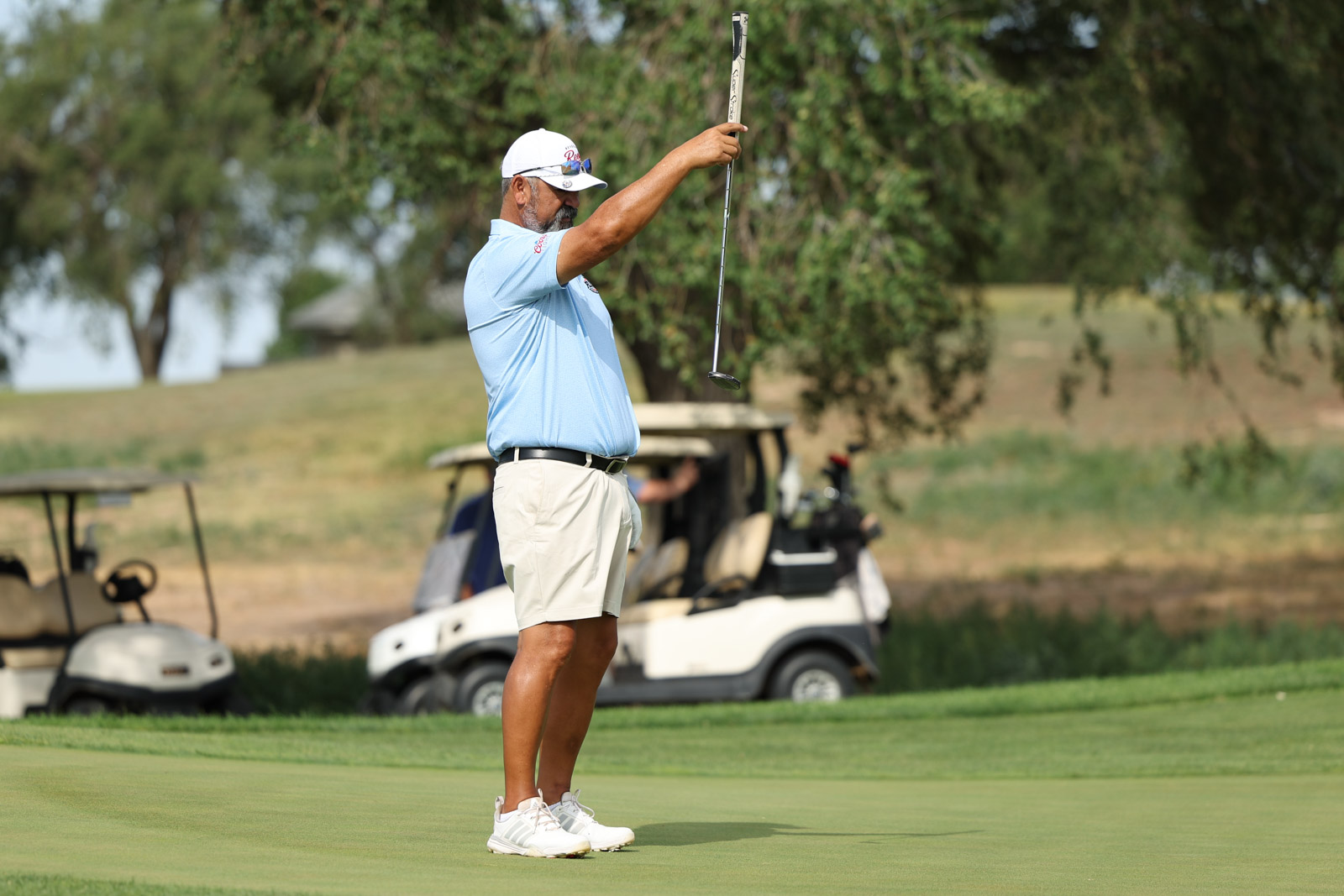 CoorTOC_FinalDay-25 | Press Pass Sports Golfer on the putting green lifts a putter overhead, wearing a light blue polo and white cap, with golf carts in the background