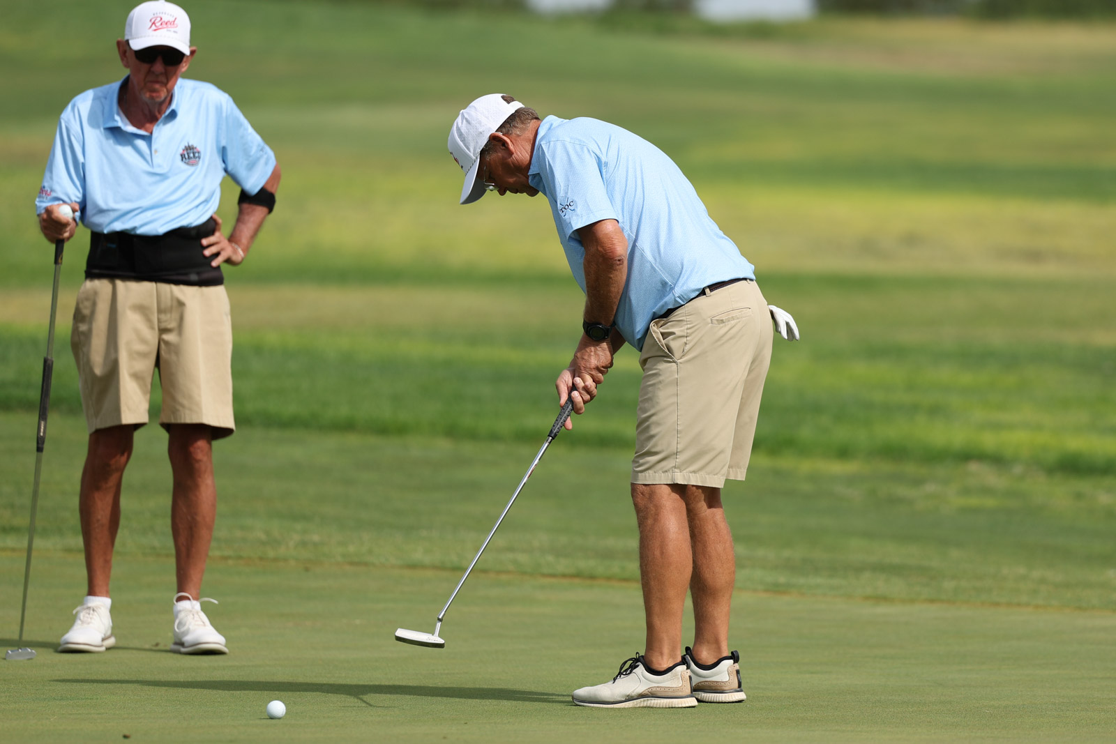 CoorTOC_FinalDay-28 | Press Pass Sports Two older men on a golf green; one puts while the other watches, holding a putter and wearing light blue shirts and khaki shorts