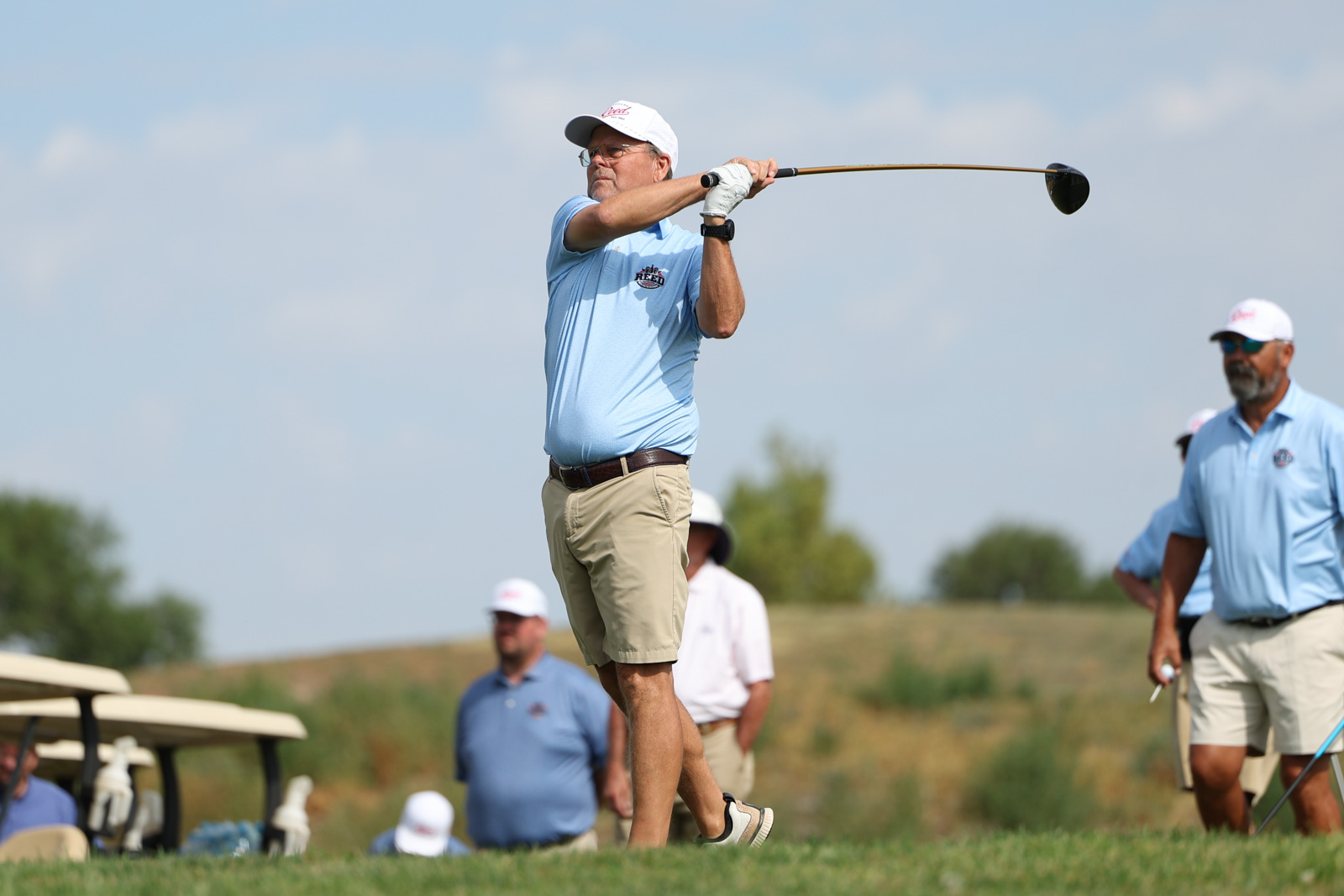 CoorTOC_FinalDay-31 | Press Pass Sports Older man in a light blue polo and beige shorts mid-swing with a golf club on a sunny course, teammates in the background.