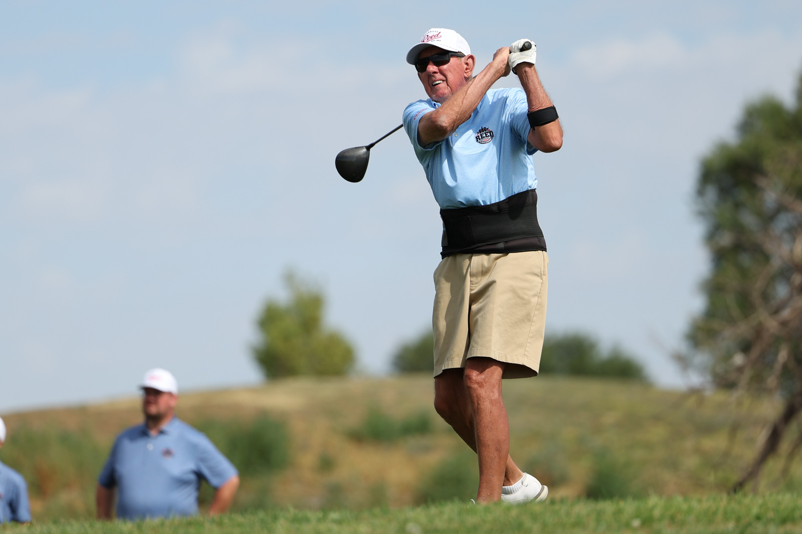 CoorTOC_FinalDay-33 | Press Pass Sports Older man in a light blue polo swings a golf club on a sunny course, wearing a white cap and beige shorts with a waist support belt.
