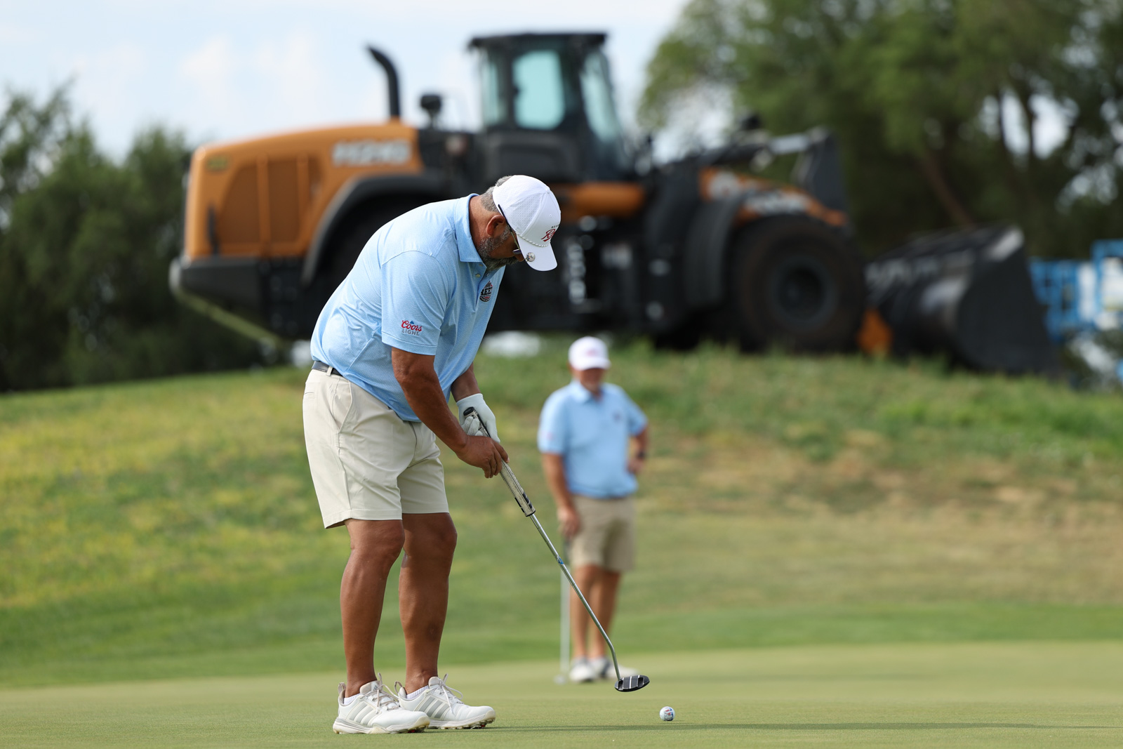 CoorTOC_FinalDay-34 | Press Pass Sports Golfer lining up a putt on a green with a large orange construction vehicle in the background and a spectator watching.