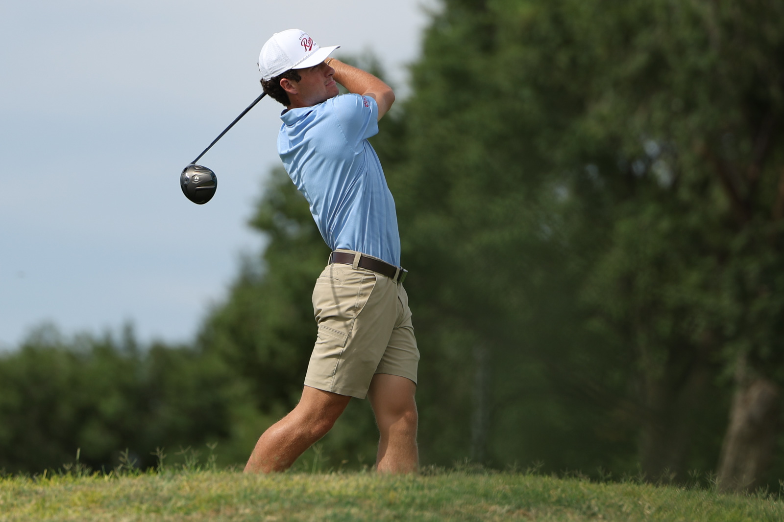 CoorTOC_FinalDay-36 | Press Pass Sports Male golfer in a light blue shirt and khaki shorts mid-swing on a sunny golf course, driver raised behind him.