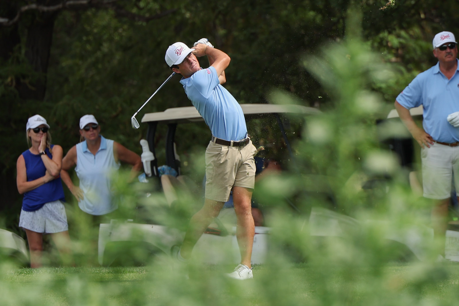 CoorTOC_FinalDay-46 | Press Pass Sports Male golfer in a light blue polo and khaki shorts mid-swing on a golf course, with spectators and a golf cart in the background.
