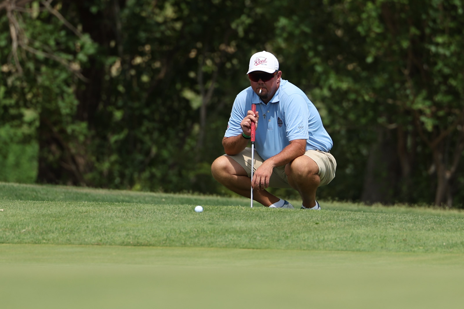 CoorTOC_FinalDay-50 | Press Pass Sports Golfer crouches on the putting green, lining up a putt with a red putter while smoking a cigarette, wearing a light blue polo and khaki shorts with a white cap and sunglasses.
