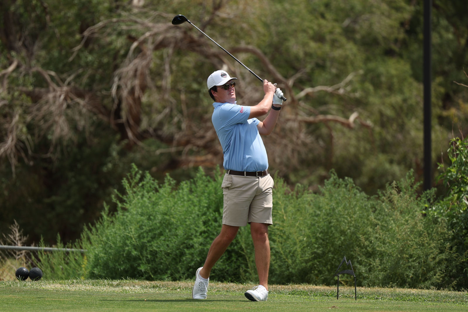 CoorTOC_FinalDay-58 | Press Pass Sports Male golfer in a light blue polo and beige shorts mid-swing on a tee, with lush greenery in the background and a white cap and sunglasses.