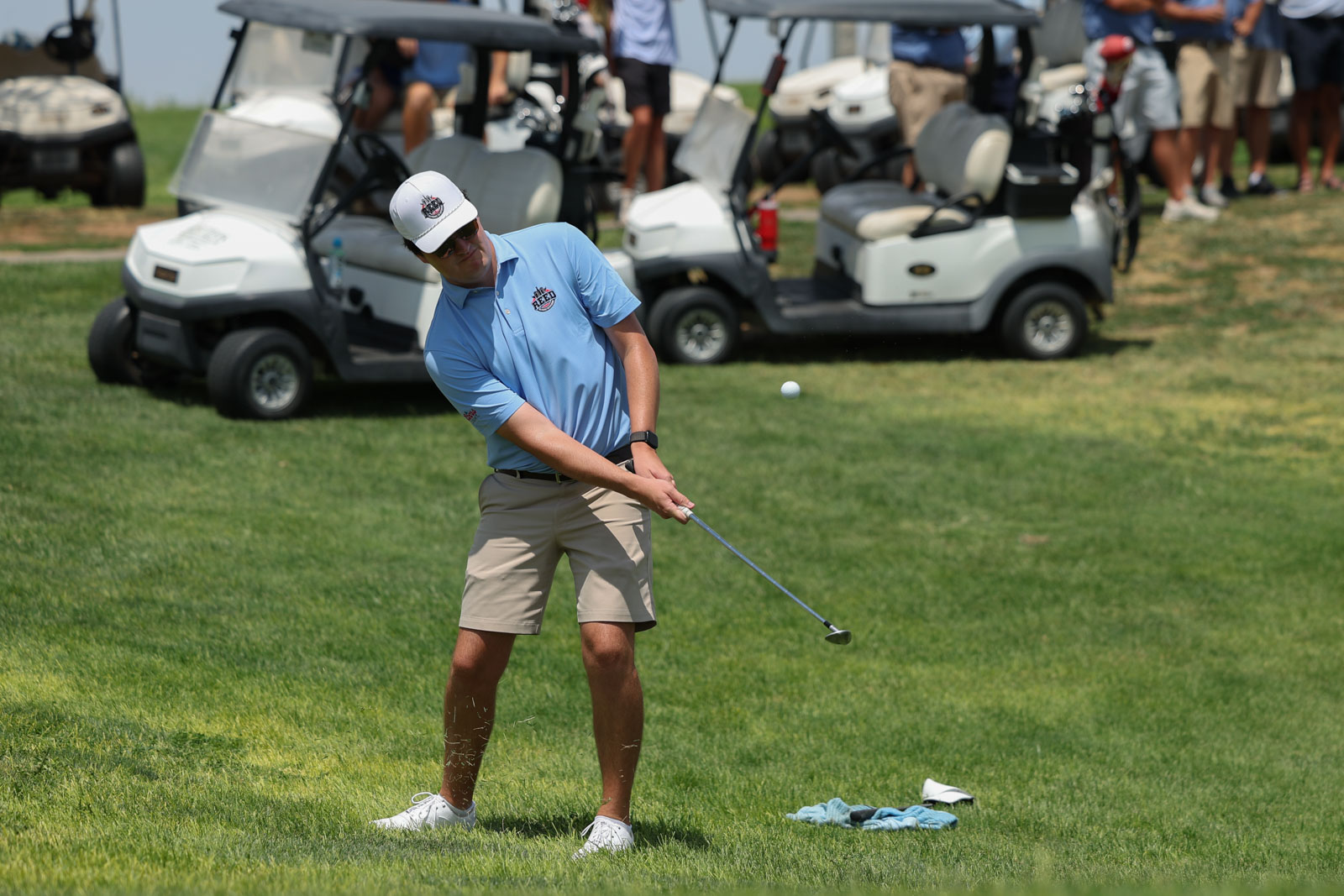 CoorTOC_FinalDay-63 | Press Pass Sports Golfer in a light blue shirt and beige shorts mid-swing on a sunny golf course, with carts and spectators in the background.