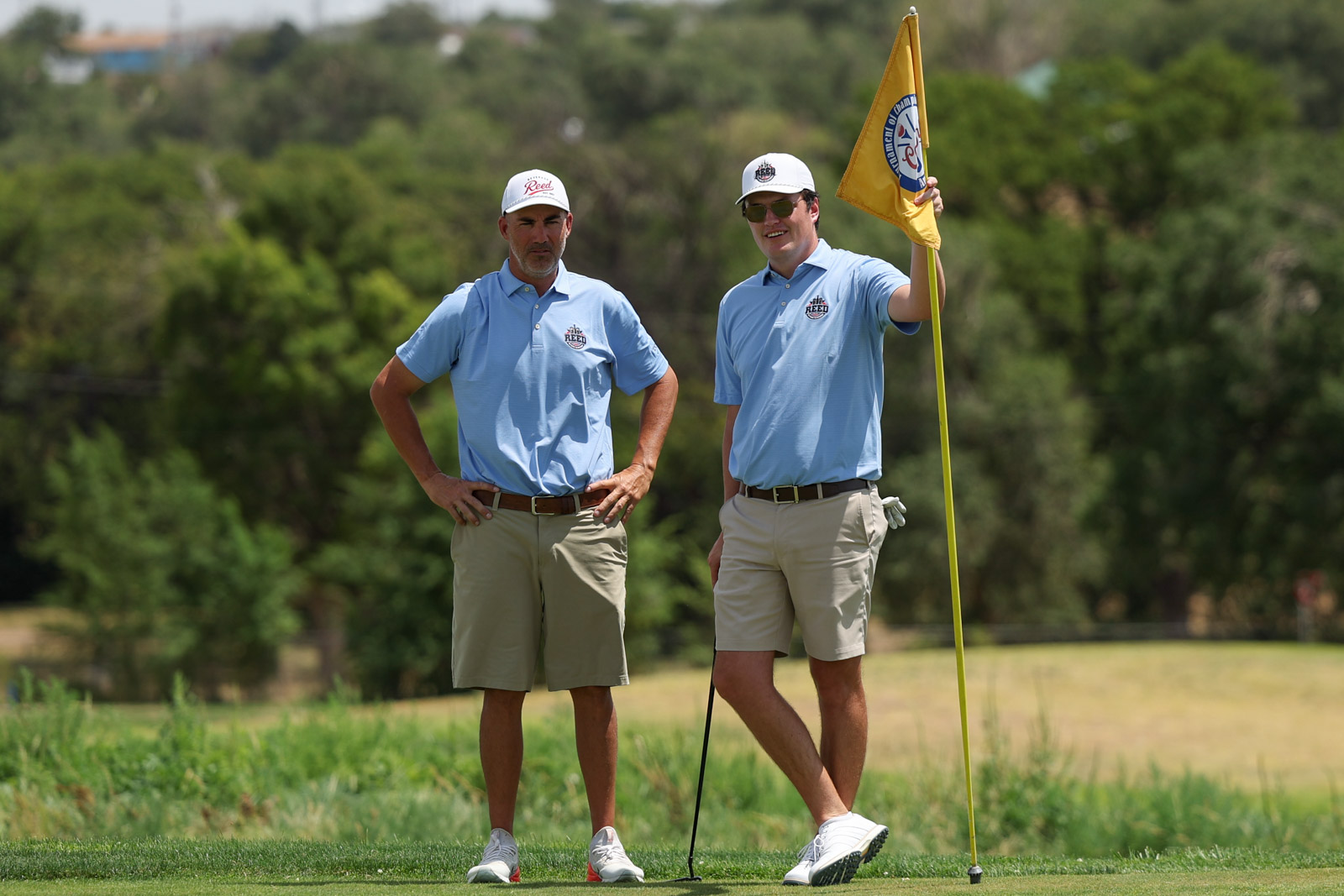 CoorTOC_FinalDay-64 | Press Pass Sports Two male golfers in light blue polo shirts stand on the green, one holding a yellow flag at the hole while the other rests his hands on his hips.