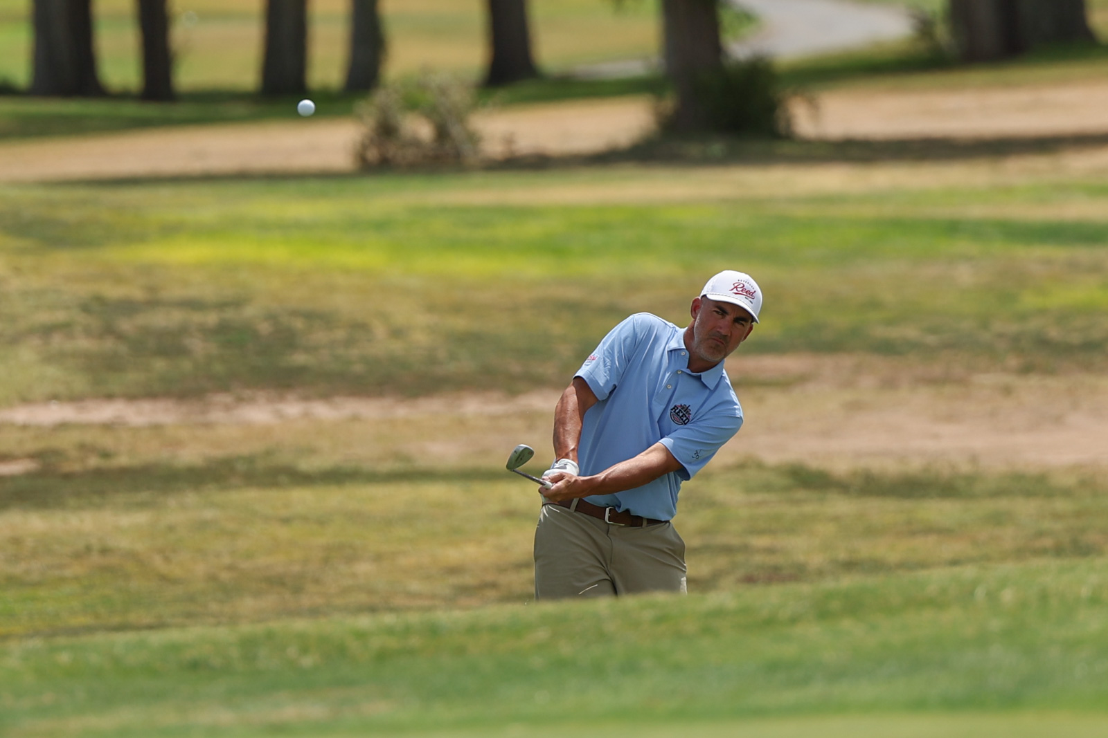 CoorTOC_FinalDay-65 | Press Pass Sports Male golfer in a light blue polo and white cap mid-swing on a grassy golf course, ball suspended in the air ahead of him