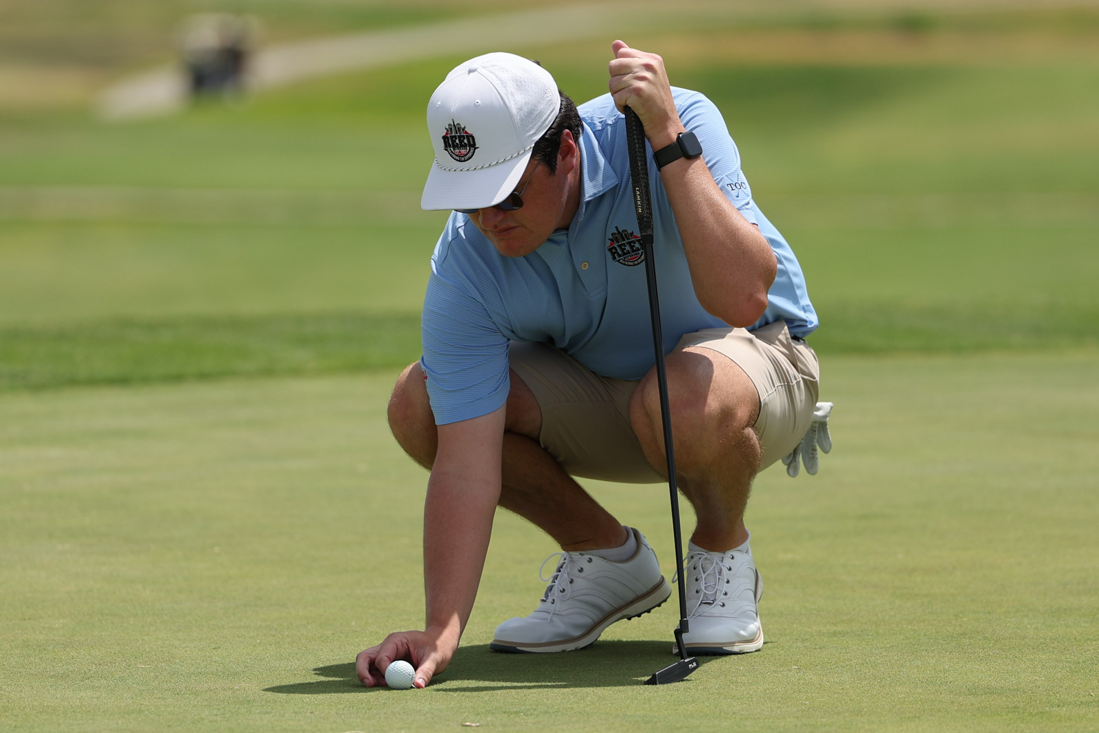 CoorTOC_FinalDay-66 | Press Pass Sports Golfer crouches on a putting green, lining up a putt with a black putter, wearing a light blue polo and white cap