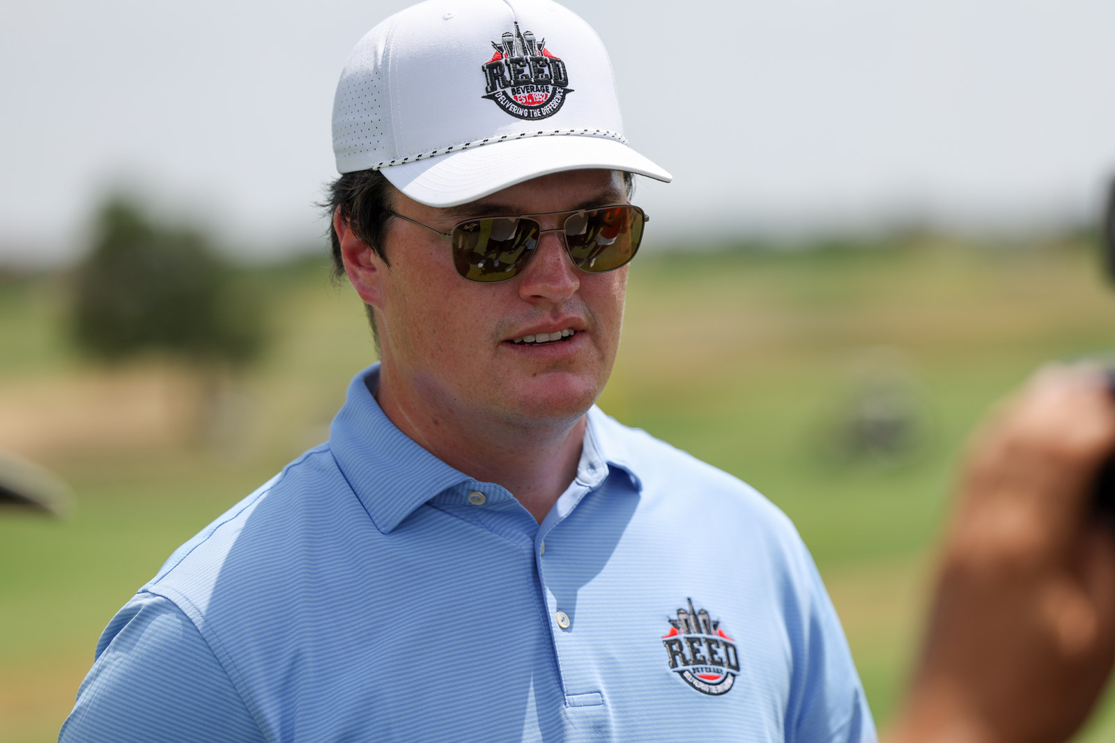 CoorTOC_FinalDay-68 | Press Pass Sports Man wearing a light blue polo with a Reed logo and a white cap, sunglasses, outdoors on a sunny day