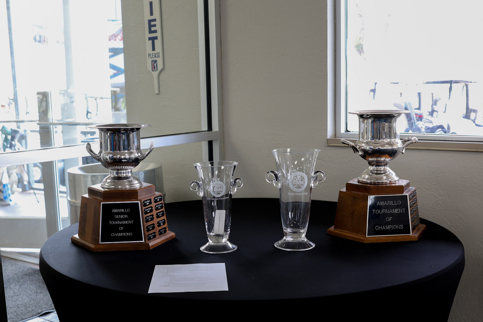 Two silver trophies on wooden bases flank etched glass championship cups on a black round table; plaques read 'Amarillo Senior Tournament of Champions.'] ,