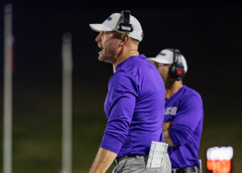 Coach in a white cap and purple shirt wearing a headset, with a clipboard tucked in his pocket during a night practice.