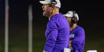 Coach in a white cap and purple shirt wearing a headset, with a clipboard tucked in his pocket during a night practice.