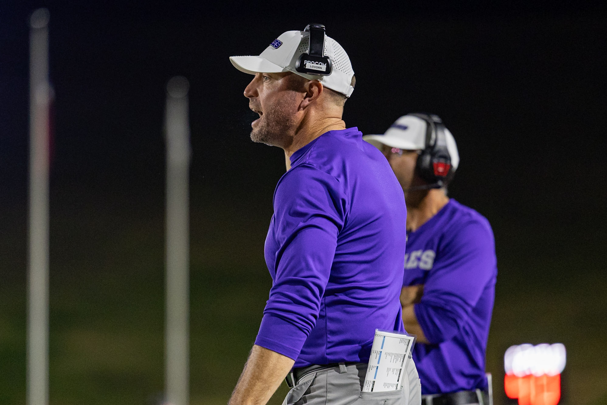 CHS-WPHS-FB-31 2 | Press Pass Sports Coach in a white cap and purple shirt wearing a headset, with a clipboard tucked in his pocket during a night practice.