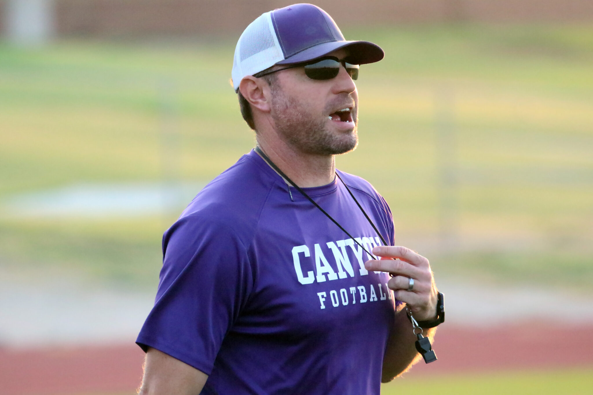 1K6A8615 | Press Pass Sports Side-profile of a man in a purple training shirt and cap, speaking with mouth open outdoors on a sports field, wearing sunglasses and a whistle/lanyard nearby.
