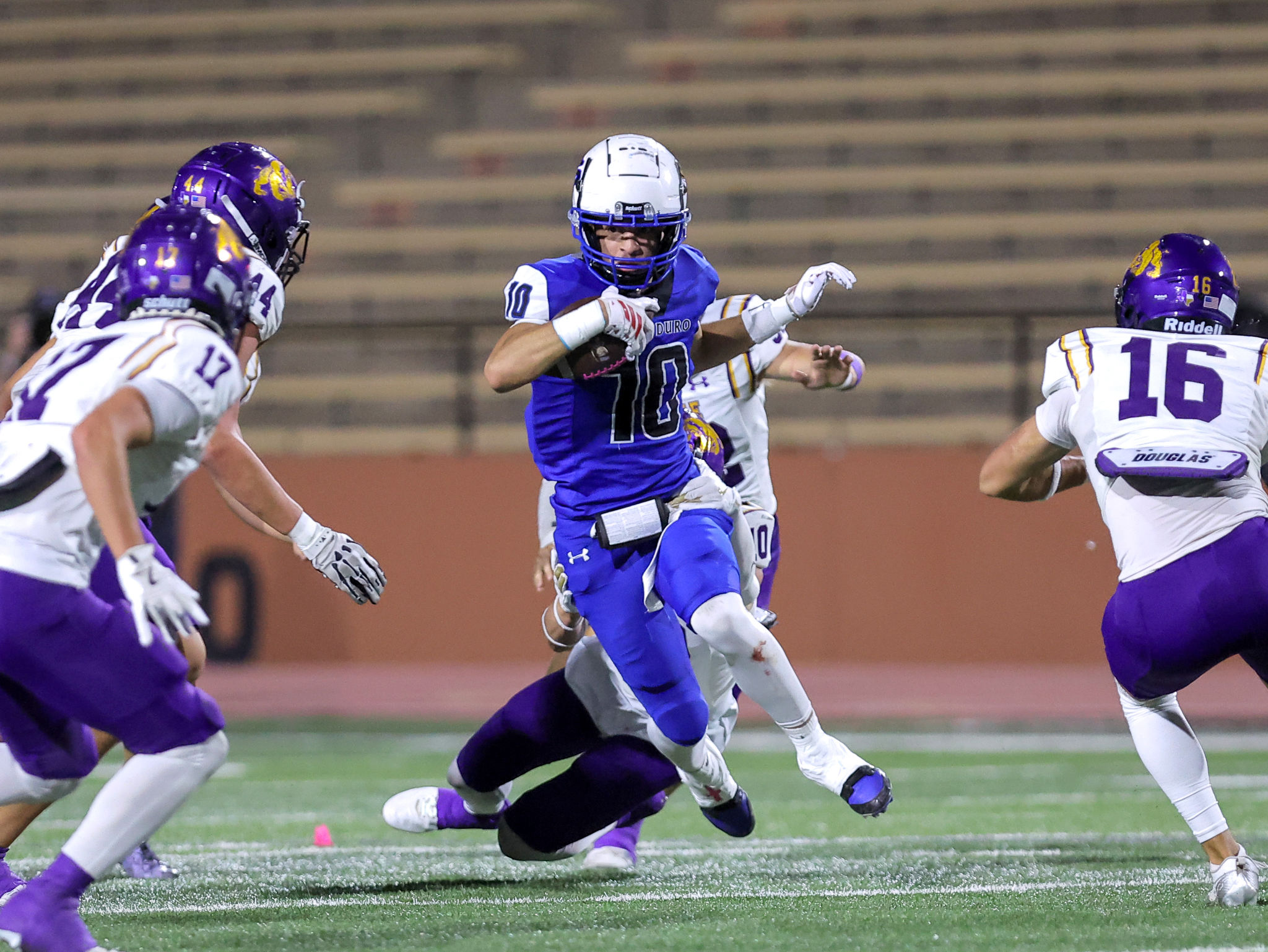 Football player in blue jersey (number 10) runs with the ball as defenders in purple and white pursue during a night game on a green field.