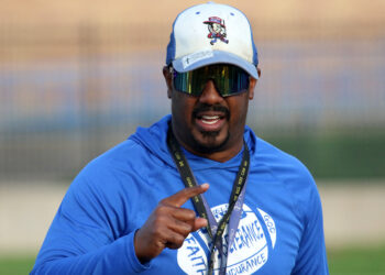 Palo Duro - Eric Mims 2 | Press Pass Sports Man in a blue athletic shirt, cap, and mirrored sunglasses smiles at the camera while gesturing with his hand during an outdoor event.