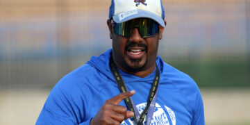 Man in a blue athletic shirt, cap, and mirrored sunglasses smiles at the camera while gesturing with his hand during an outdoor event.