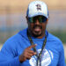 Palo Duro - Eric Mims 2 | Press Pass Sports Man in a blue athletic shirt, cap, and mirrored sunglasses smiles at the camera while gesturing with his hand during an outdoor event.