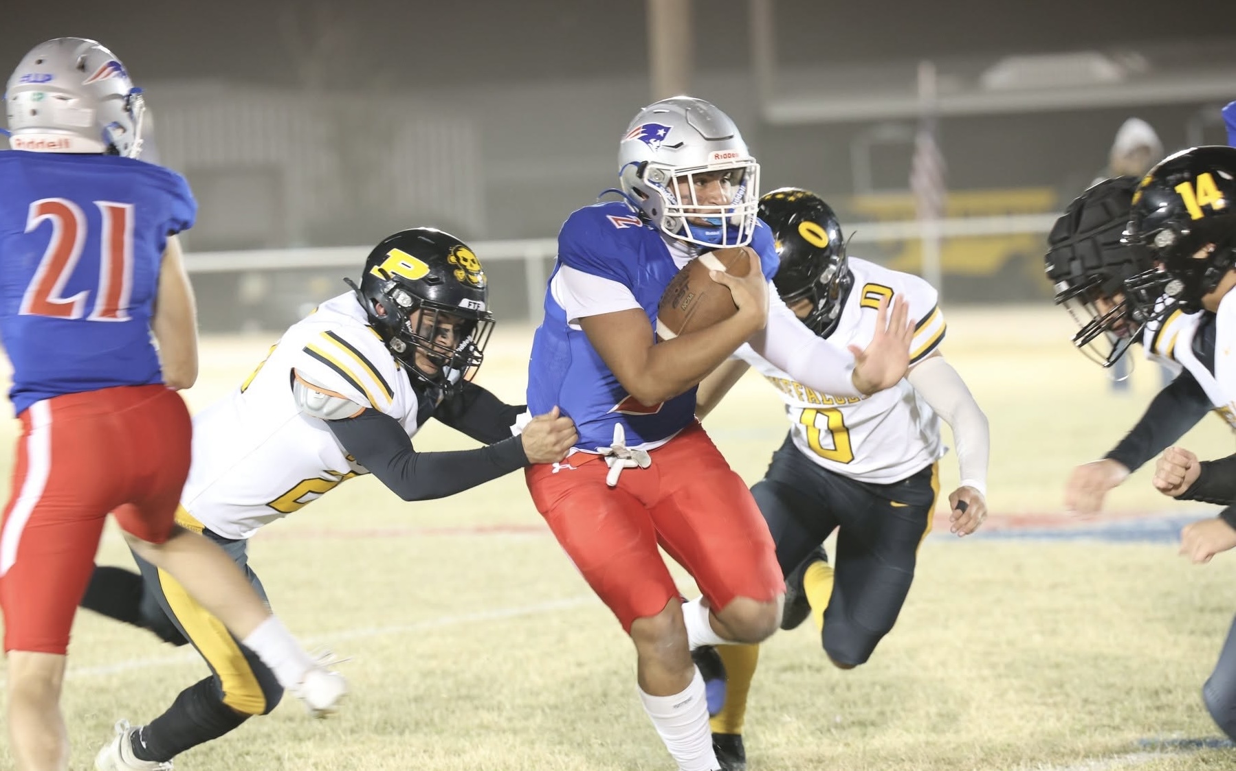 A football runner in a blue jersey hugs the ball while defenders in white and yellow close in on him on a grassy field.