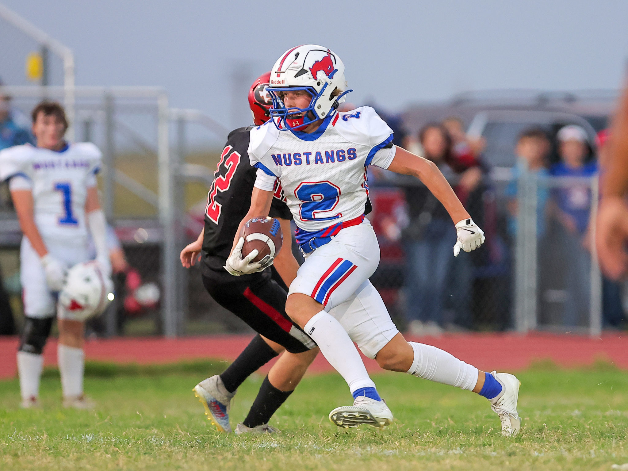 Male football player in a white Mustang jersey runs with the ball during a game, pursued by a defender in black.