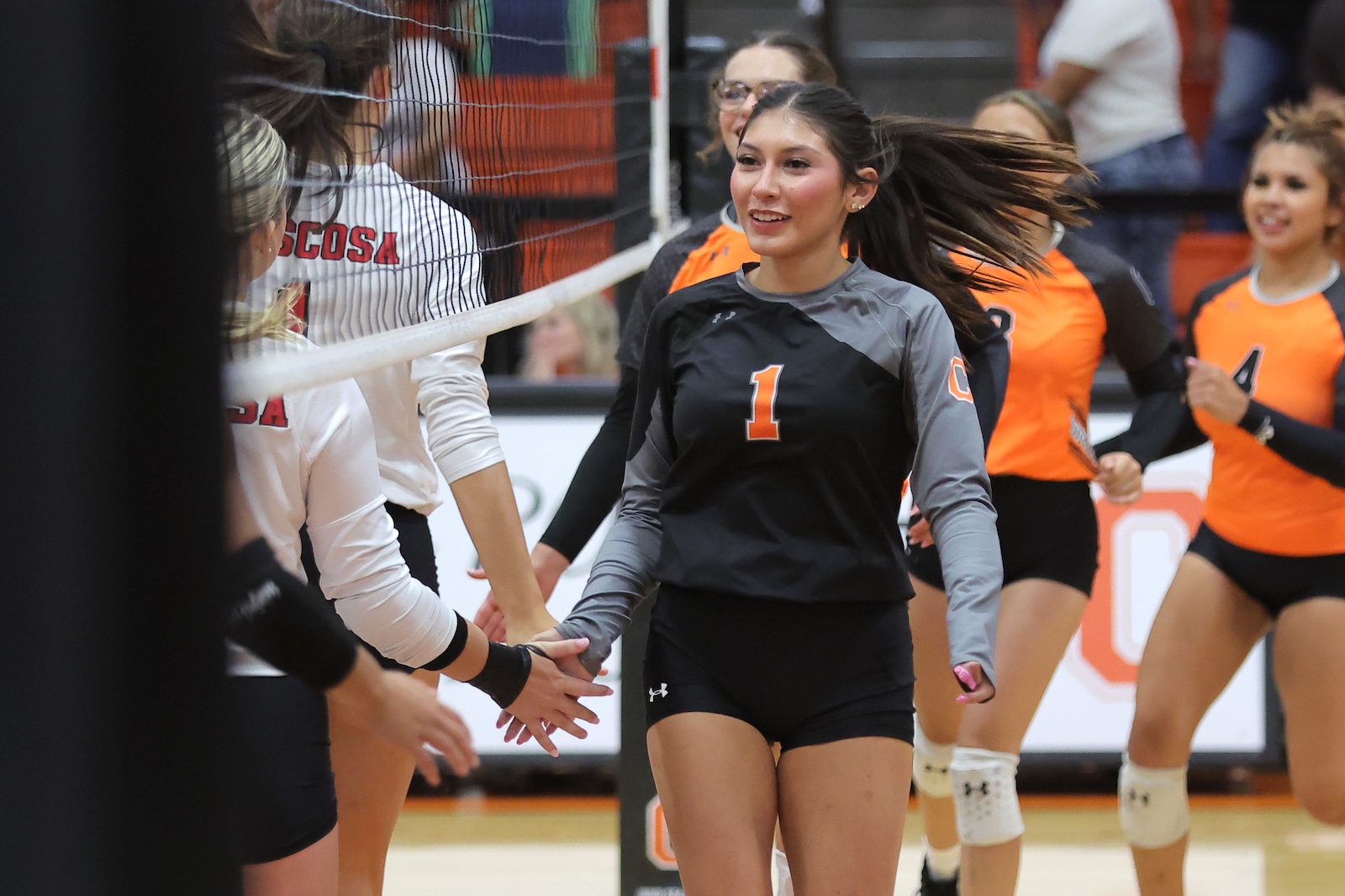 Smiling female volleyball players in black, orange, and white uniforms high-five at the net after a match in a gym.