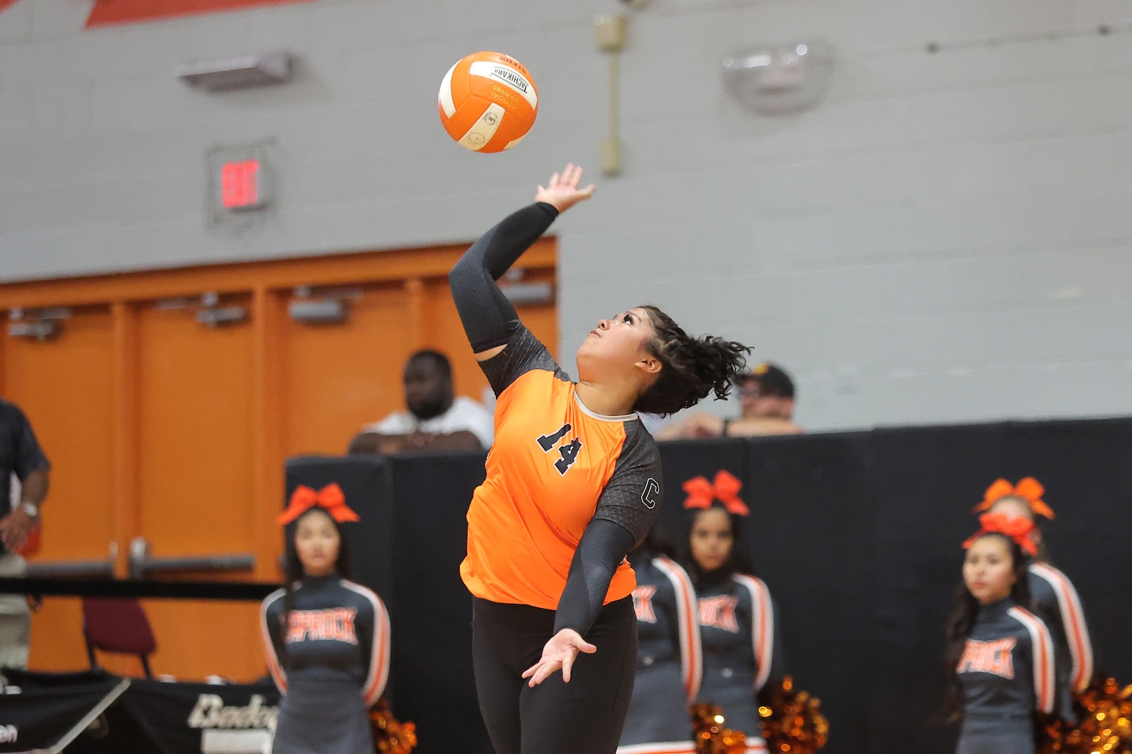 Female volleyball player in an orange jersey serves the ball during a match in a gym, with teammates cheering in the background.