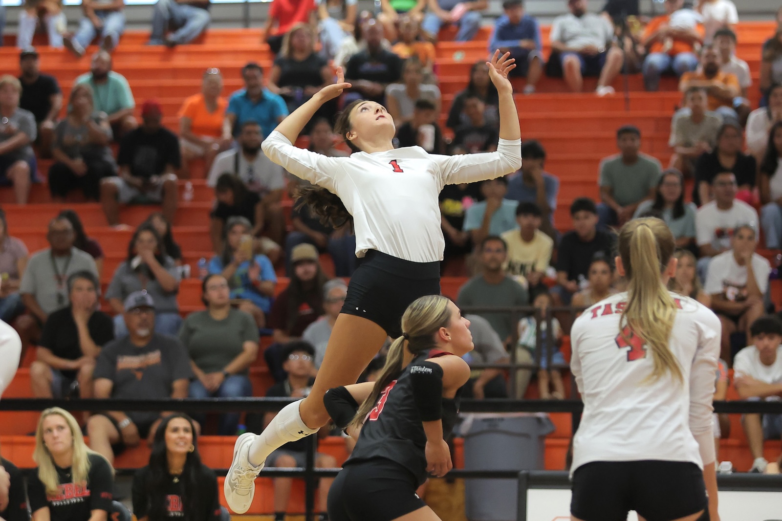 Female volleyball player in white jersey (number 1) leaps to spike the ball, with orange bleachers and spectators in the background.