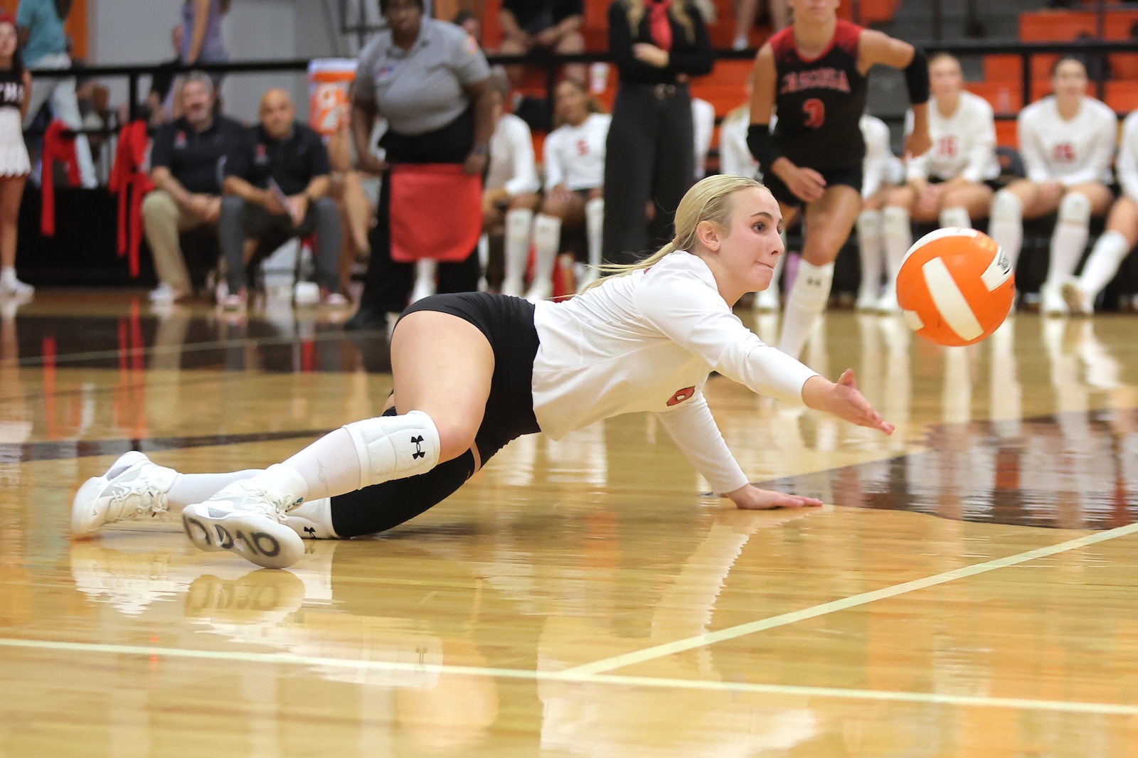 Female volleyball player diving on a polished gym floor to keep the ball in play, white jersey and knee pads visible, orange volleyball near her hand.