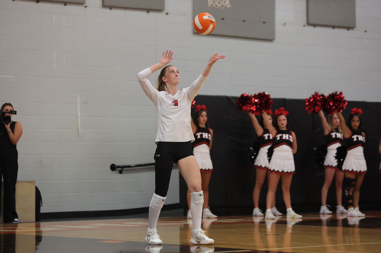 Female volleyball player in white jersey with number 1 serves the ball on a gym court, teammates and cheerleaders with red pom-poms in the background.