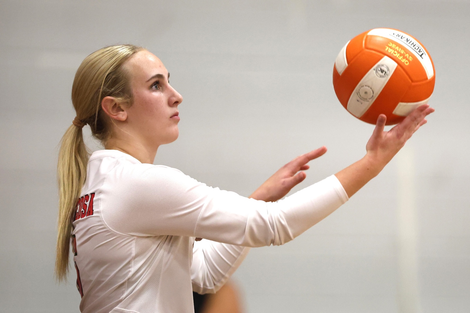 Female volleyball player in a white jersey preparing to set a bright orange volleyball during practice.